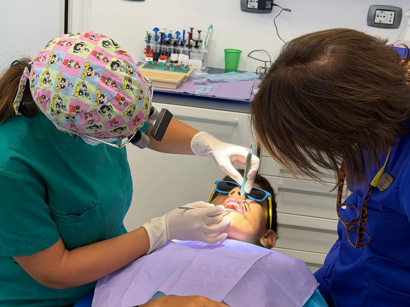 Bambino con gli occhiali da sole colorati sulla poltrona mentre la Dott.ssa Giorgia conta i dentini con lo specchietto